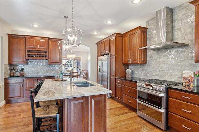 kitchen with light wood finished floors, dark stone countertops, appliances with stainless steel finishes, wall chimney exhaust hood, and a sink