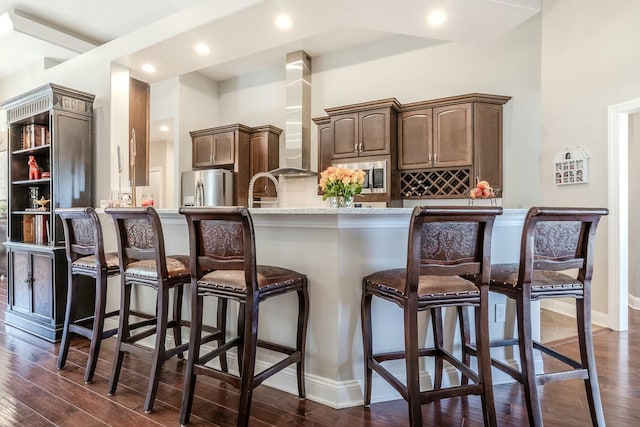 kitchen with wall chimney exhaust hood, light countertops, appliances with stainless steel finishes, dark wood-type flooring, and a kitchen breakfast bar