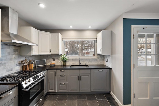 kitchen featuring stainless steel gas stove, dark countertops, wall chimney range hood, white cabinetry, and a sink