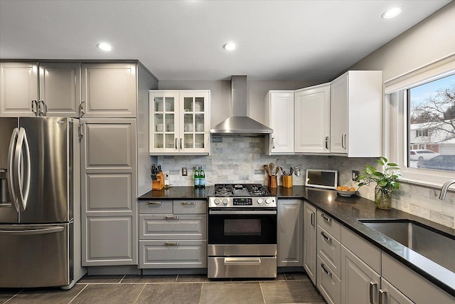 kitchen featuring wall chimney exhaust hood, glass insert cabinets, a sink, stainless steel appliances, and backsplash