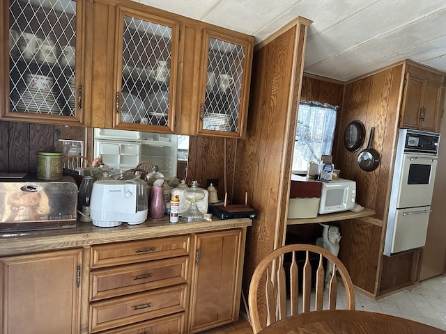 kitchen featuring wooden walls, white appliances, light countertops, brown cabinetry, and glass insert cabinets