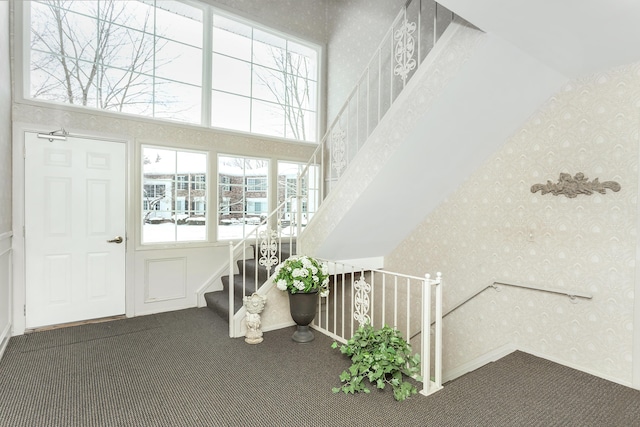 entryway with stairs, dark colored carpet, a towering ceiling, and wallpapered walls