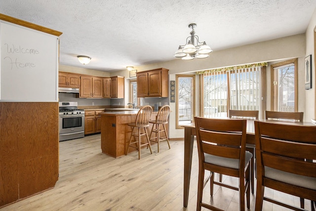 kitchen featuring stainless steel range with gas cooktop, decorative light fixtures, brown cabinetry, under cabinet range hood, and a kitchen breakfast bar