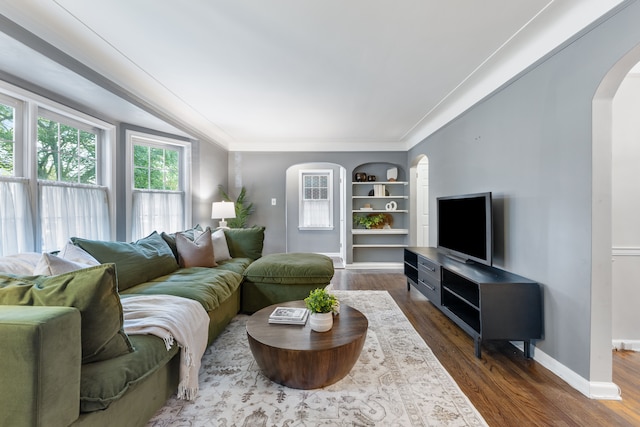 living room featuring baseboards, arched walkways, dark wood-type flooring, crown molding, and built in shelves