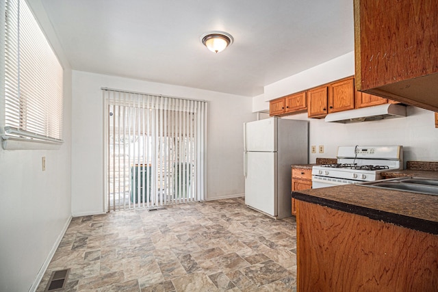 kitchen with brown cabinets, dark countertops, white appliances, under cabinet range hood, and baseboards