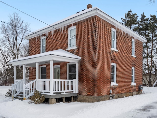 snow covered property with covered porch, a chimney, and brick siding