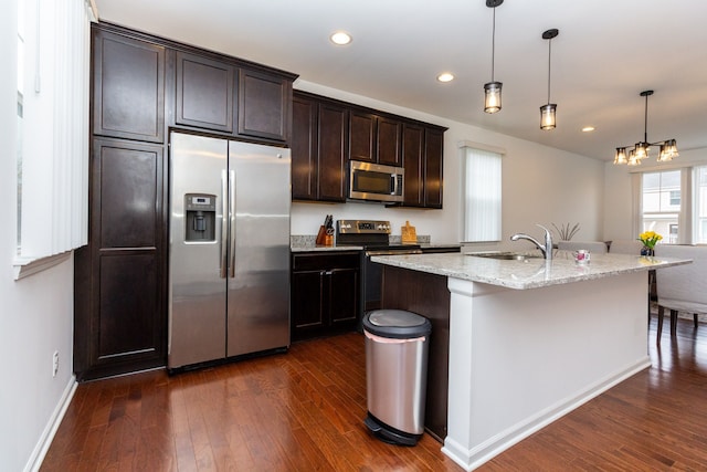 kitchen featuring a kitchen island with sink, stainless steel appliances, a sink, light stone countertops, and decorative light fixtures