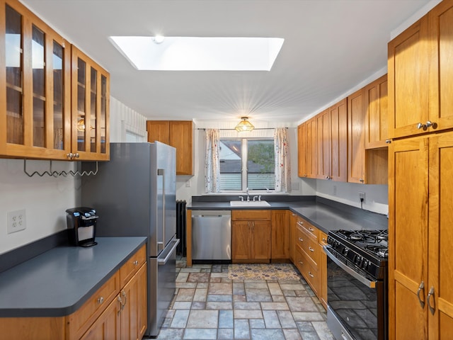 kitchen featuring stainless steel appliances, a skylight, brown cabinets, dark countertops, and glass insert cabinets