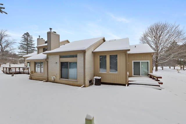 snow covered back of property featuring a deck, brick siding, and a chimney