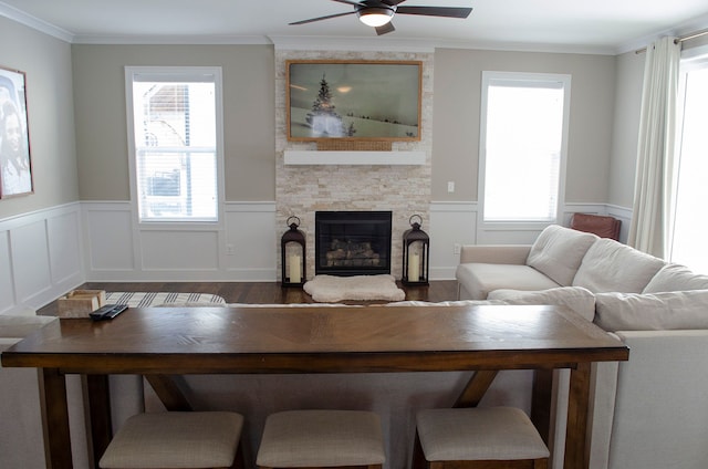 living room with a ceiling fan, wainscoting, ornamental molding, wood finished floors, and a stone fireplace