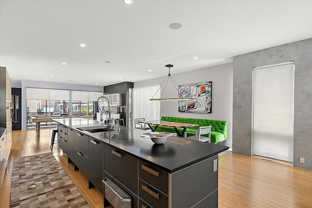 kitchen featuring pendant lighting, dark countertops, light wood-style flooring, a sink, and an island with sink