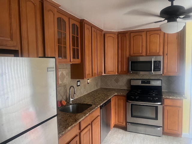 kitchen featuring appliances with stainless steel finishes, brown cabinetry, glass insert cabinets, a sink, and dark stone countertops