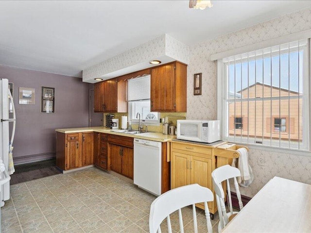 kitchen featuring light countertops, brown cabinetry, a sink, white appliances, and wallpapered walls