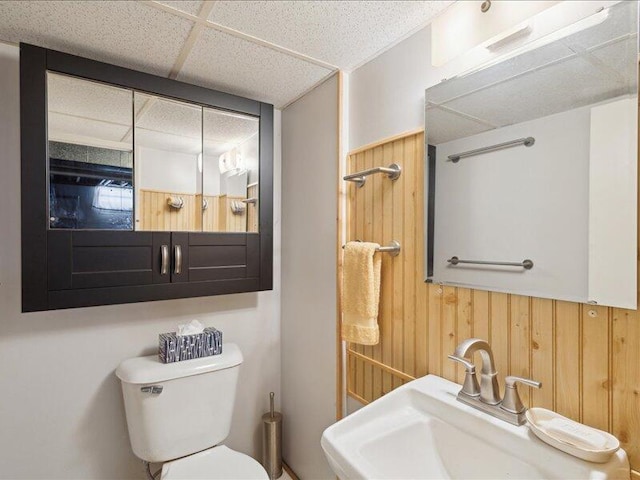 bathroom with a paneled ceiling, wood walls, toilet, and a sink