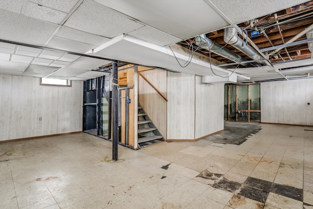 basement featuring light floors, stairs, baseboards, and wood walls