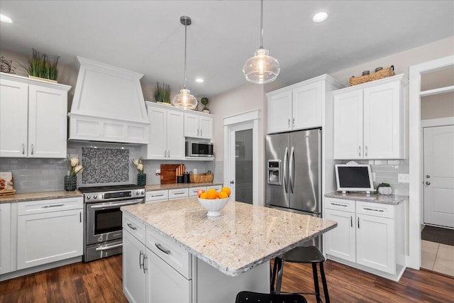 kitchen featuring white cabinets, appliances with stainless steel finishes, a center island, decorative light fixtures, and premium range hood