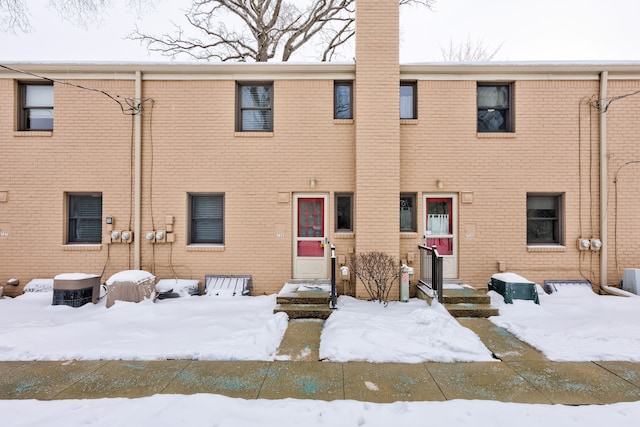 snow covered back of property with entry steps, brick siding, and a chimney