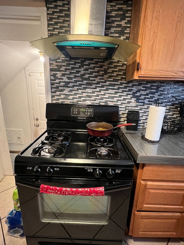 kitchen featuring brown cabinetry, decorative backsplash, black gas range oven, dark countertops, and wall chimney range hood