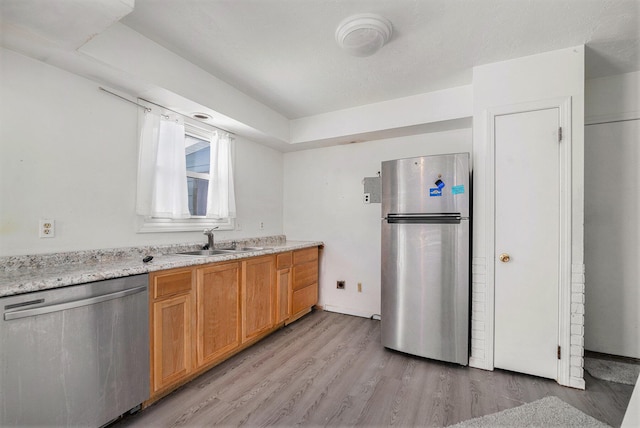 kitchen featuring light wood finished floors, brown cabinetry, appliances with stainless steel finishes, light stone countertops, and a sink