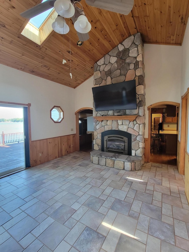 unfurnished living room with a skylight, wainscoting, wood ceiling, a stone fireplace, and high vaulted ceiling