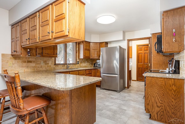 kitchen featuring black microwave, a peninsula, a sink, freestanding refrigerator, and brown cabinetry