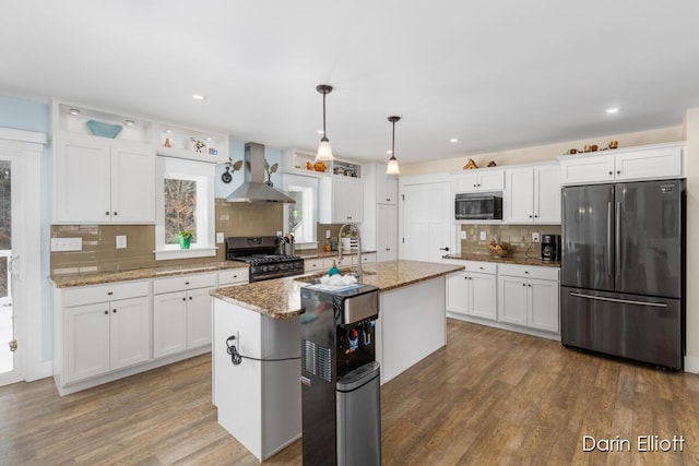 kitchen featuring pendant lighting, appliances with stainless steel finishes, a kitchen island with sink, white cabinetry, and wall chimney range hood