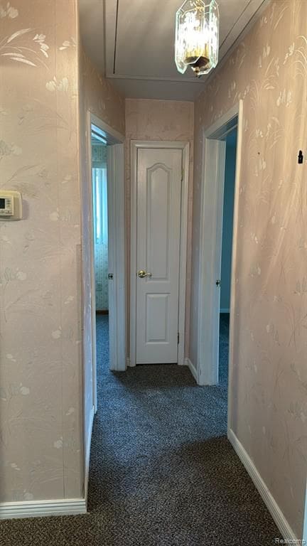 hallway featuring wallpapered walls, attic access, dark carpet, and baseboards