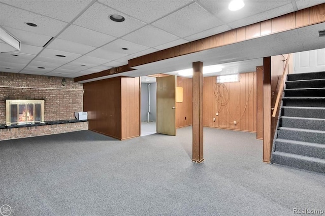 below grade area with wood walls, stairway, a brick fireplace, and a drop ceiling