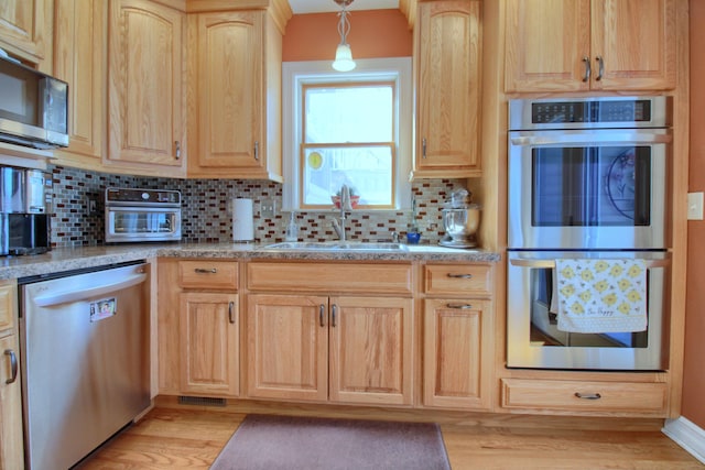 kitchen featuring light stone counters, decorative light fixtures, stainless steel appliances, light brown cabinetry, and a sink