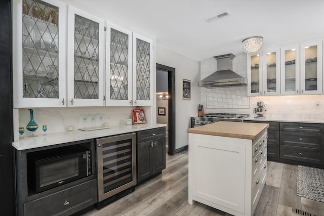 kitchen with wine cooler, white cabinetry, wall chimney range hood, dark cabinetry, and glass insert cabinets