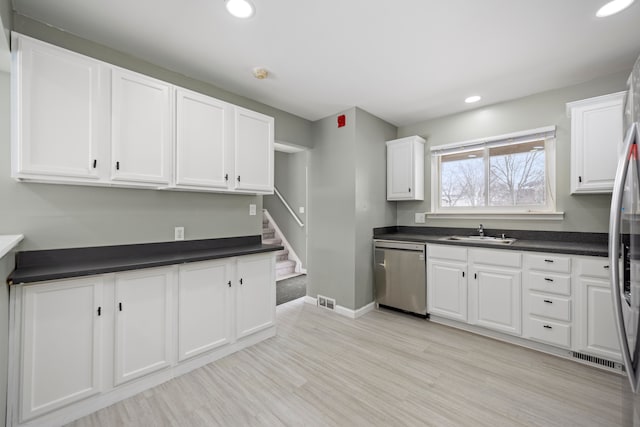 kitchen featuring light wood finished floors, dark countertops, stainless steel dishwasher, white cabinets, and a sink