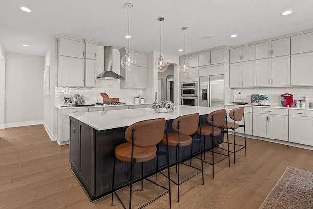 kitchen featuring decorative light fixtures, light countertops, wall chimney range hood, appliances with stainless steel finishes, and a center island with sink