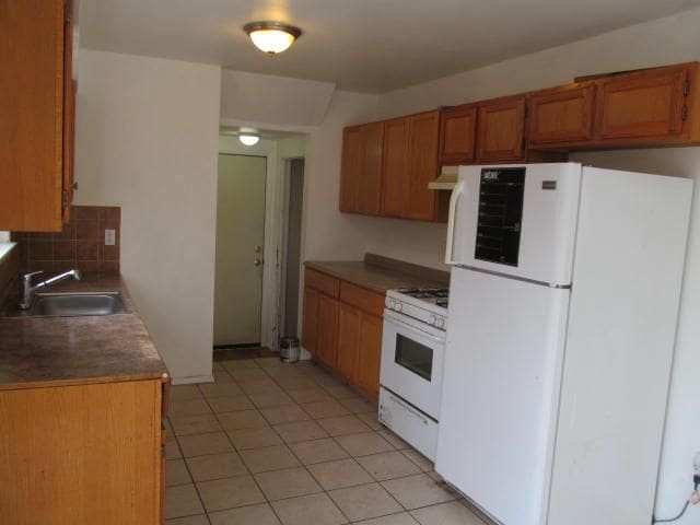 kitchen featuring white appliances, light tile patterned floors, decorative backsplash, brown cabinetry, and a sink