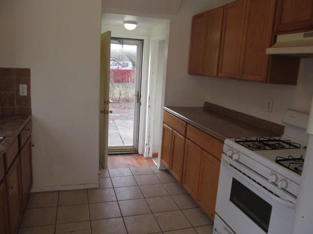 kitchen featuring dark countertops, brown cabinets, white gas range, under cabinet range hood, and light tile patterned flooring