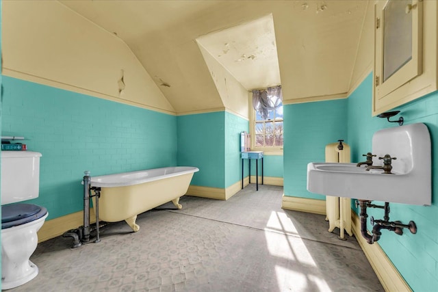 bathroom with a freestanding bath, toilet, vaulted ceiling, brick wall, and baseboards