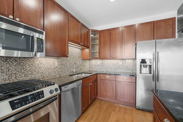 kitchen featuring light wood-style flooring, a sink, appliances with stainless steel finishes, dark stone counters, and glass insert cabinets