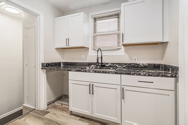 kitchen featuring light wood-style flooring, white cabinets, a sink, dark stone counters, and baseboards