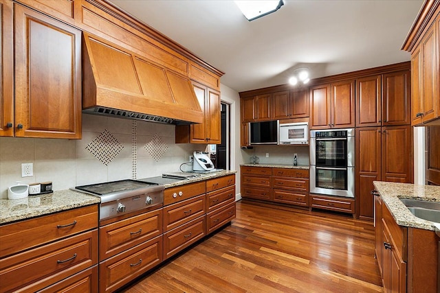 kitchen with appliances with stainless steel finishes, brown cabinetry, light stone counters, and wood finished floors