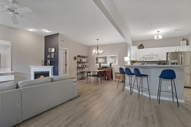 living room with visible vents, light wood-style flooring, a warm lit fireplace, beamed ceiling, and ceiling fan with notable chandelier