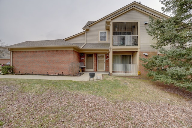 rear view of property featuring brick siding, a balcony, a shingled roof, and a yard