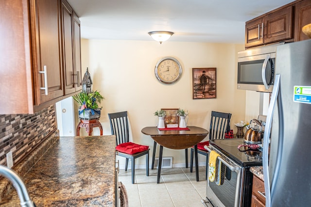 kitchen featuring light tile patterned flooring, appliances with stainless steel finishes, tasteful backsplash, brown cabinetry, and dark stone countertops