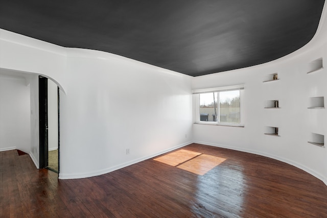 empty room featuring arched walkways, dark wood finished floors, and baseboards