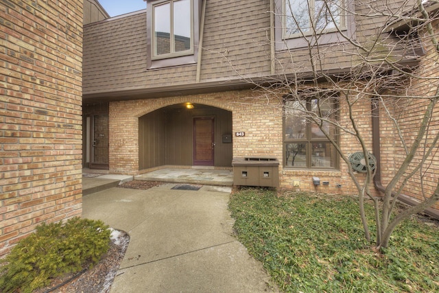 doorway to property featuring a patio, a shingled roof, mansard roof, and brick siding