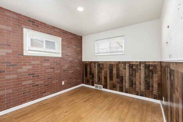 unfurnished room featuring brick wall, hardwood / wood-style floors, wainscoting, and visible vents