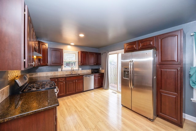 kitchen featuring stainless steel appliances, a sink, light wood-type flooring, dark stone counters, and under cabinet range hood