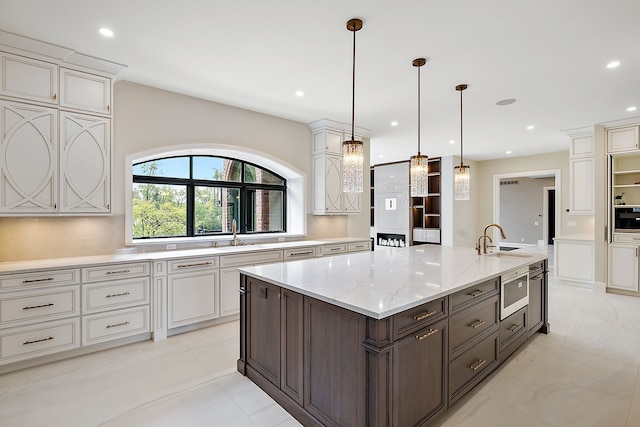 kitchen with an island with sink, light stone counters, hanging light fixtures, dark brown cabinets, and a sink