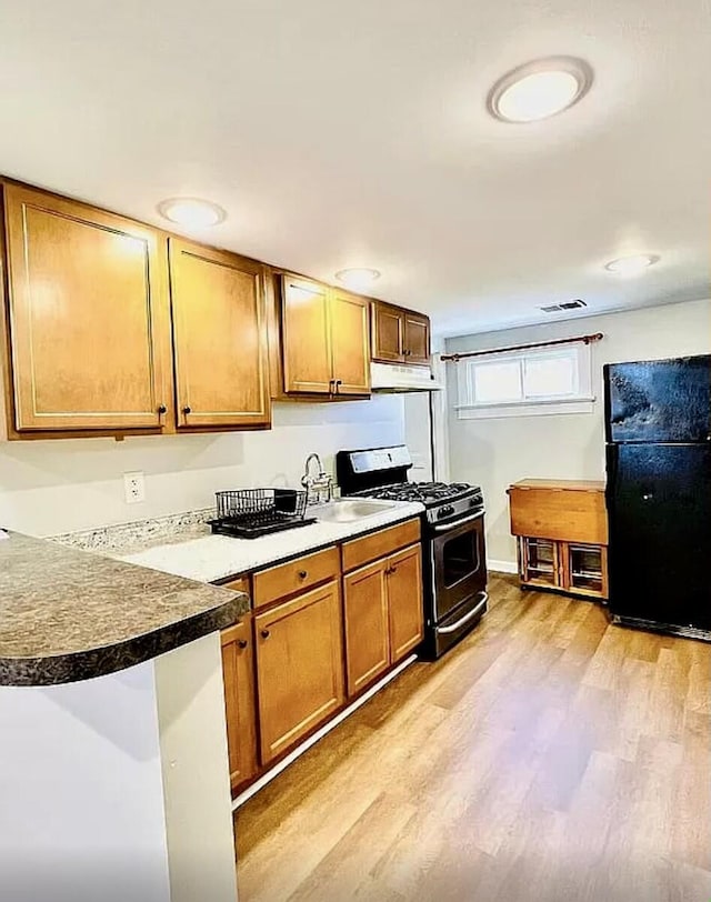 kitchen with under cabinet range hood, stainless steel gas range oven, brown cabinets, and freestanding refrigerator