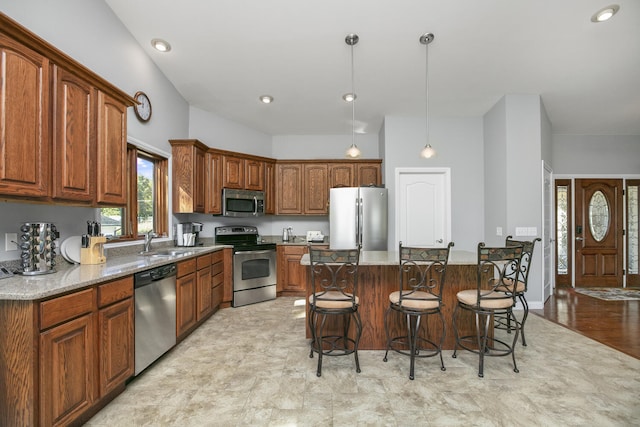 kitchen with brown cabinetry, appliances with stainless steel finishes, hanging light fixtures, light stone countertops, and a sink