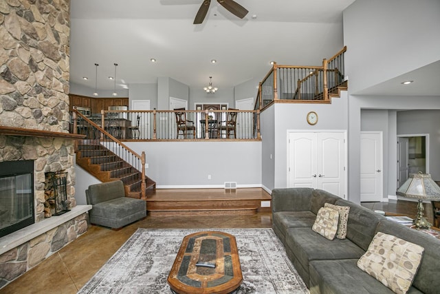 living room featuring stairs, visible vents, a towering ceiling, a stone fireplace, and ceiling fan with notable chandelier