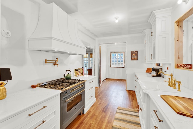 kitchen with custom exhaust hood, white cabinetry, light stone countertops, light wood-type flooring, and high end stove